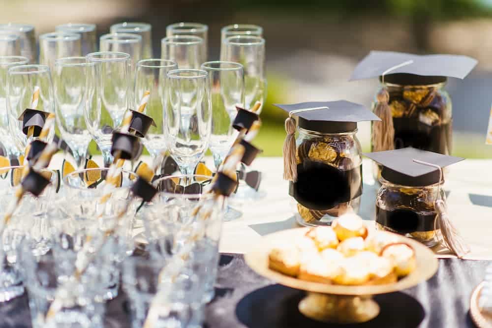 A graduation party table display featuring champagne flutes with mortarboard drink stirrers, glass jars filled with treats topped with mini graduation caps, and a gold tray of decorated cookies.